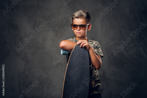 Young schoolboy in sunglasses is posing at dark photo studio with his skateboard. photo