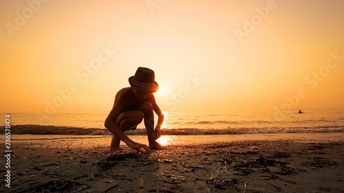 Boy with hat seeking a flat stones on beach to throw them as skipping stones on sea water in Slow Motion, cinematic sunset sun shining