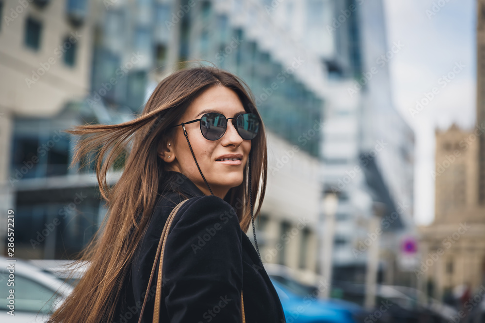 Young beautiful stylish girl in Sunglasses walking on street. Close up portrait of woman is turn around at camera and smiles. Female playfully looks at the camera. Young beautiful stylish girl in Sunglasses walking on street. Close up portrait of woman is turn around at camera and smiles. Female playfully looks at the camera.