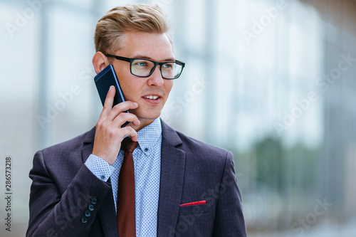 Young handsome Caucasian businessman in glasses talking on mobile phone in front of office building. Male executive manager in trendy clothes using smartphone outdoors. Modern technology concept