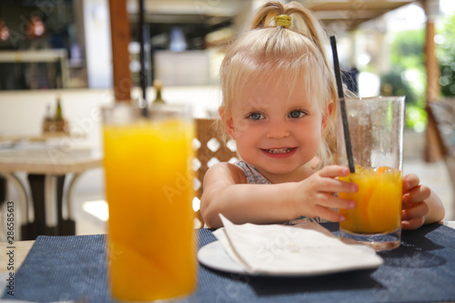 Happy toddler girl sitting at the restaurant table and drinking fresh-squeezed orange juice