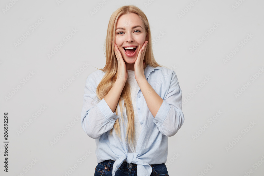 Happy pretty young lady with long blond hair wearing casual clothes, looking surprised and joyful, holding palms on her cheeks, isolated over white background Happy pretty young lady with long blond hair wearing casual clothes, looking surprised and joyful, holding palms on her cheeks, isolated over white background
