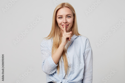 Horizontal shot of beautiful blonde long haired female keeping finger on smiling lips, demonstrating hush sign, asking to keep silence, standing over white background