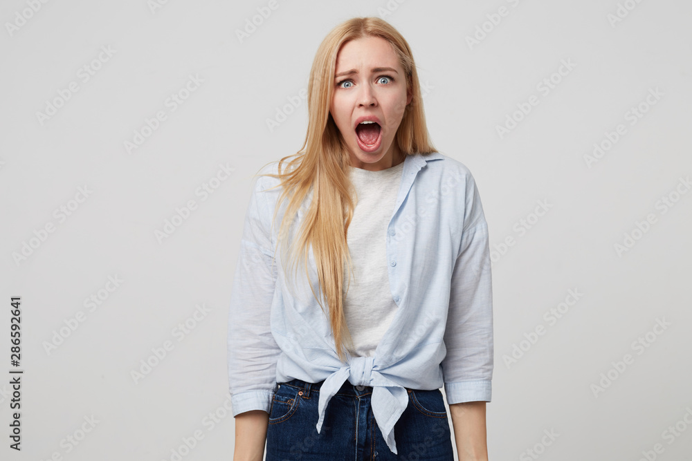 Shocked attractive young blonde long haired woman standing over white background with hands down, looking amazed to camera with wide opened mouth Shocked attractive young blonde long haired woman standing over white background with hands down, looking amazed to camera with wide opened mouth