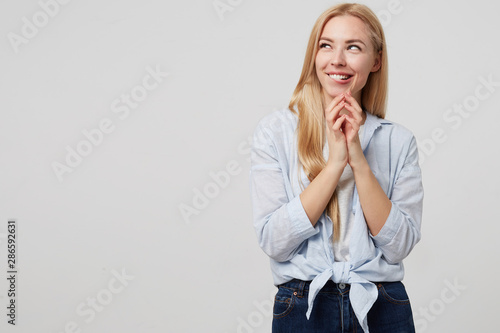 Studio shot of attractive young woman in blue shirt and jeans smiling slyly and looking away, squinting and biting underlip with folded palms, isolated over white background