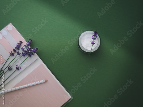 Face cream made from lavender and pink notebook with pencil on green background. Overhead shot. photo