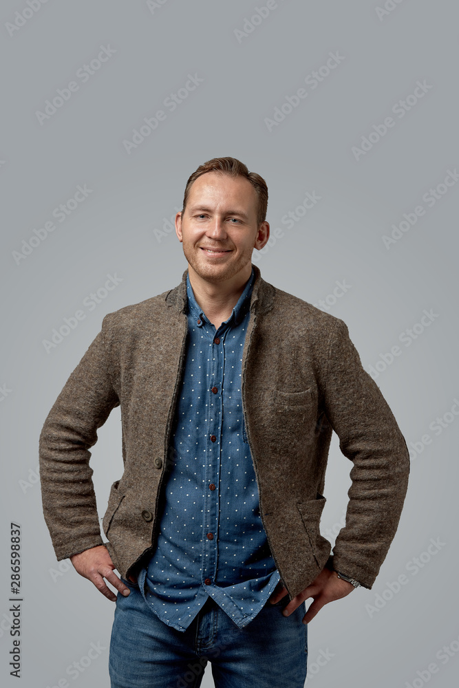 Fototapeta premium smiling old casual man in leather jacket with hands in pockets standing against gray studio wall