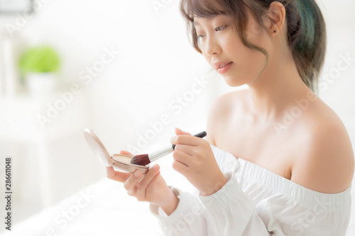 Beauty portrait young asian woman smiling with face looking mirror applying makeup with brush cheek in the bedroom, beautiful girl holding blusher facial, skin care and cosmetic concept.