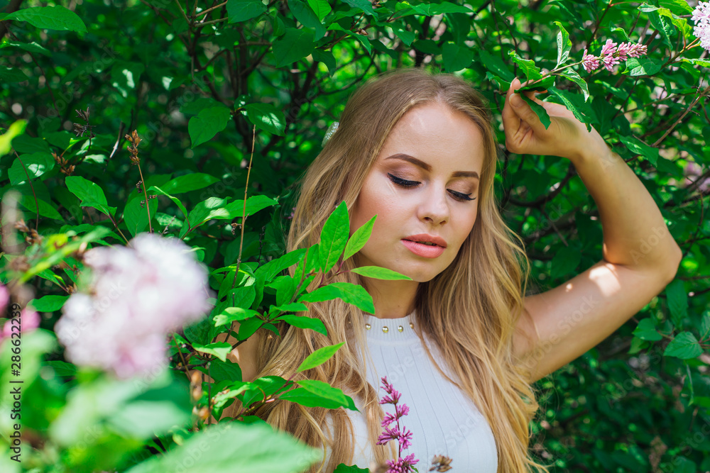 Portrait of a charming blond woman wearing beautiful white dress standing next to lilac bush. Portrait of a charming blond woman wearing beautiful white dress standing next to lilac bush.