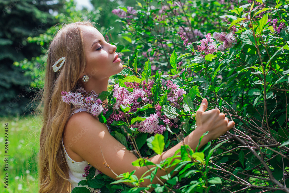Fototapeta premium Portrait of a charming blond woman wearing beautiful white dress standing next to lilac bush.