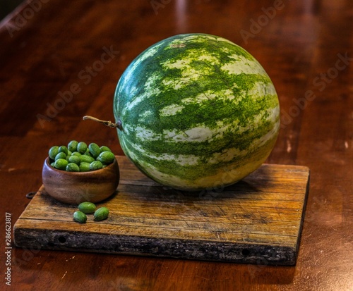 Still Life of a Large Watermelon on a Wooden Board next to a small wooden bowl of Mexican Sour Gherkins, selective focus on foreground