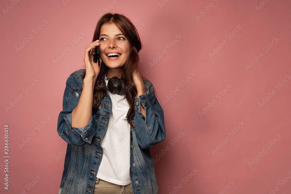 Brunette on a pink background in jeans with headphones around her neck happily smiling on the phone. Brunette on a pink background in jeans with headphones around her neck happily smiling on the phone.