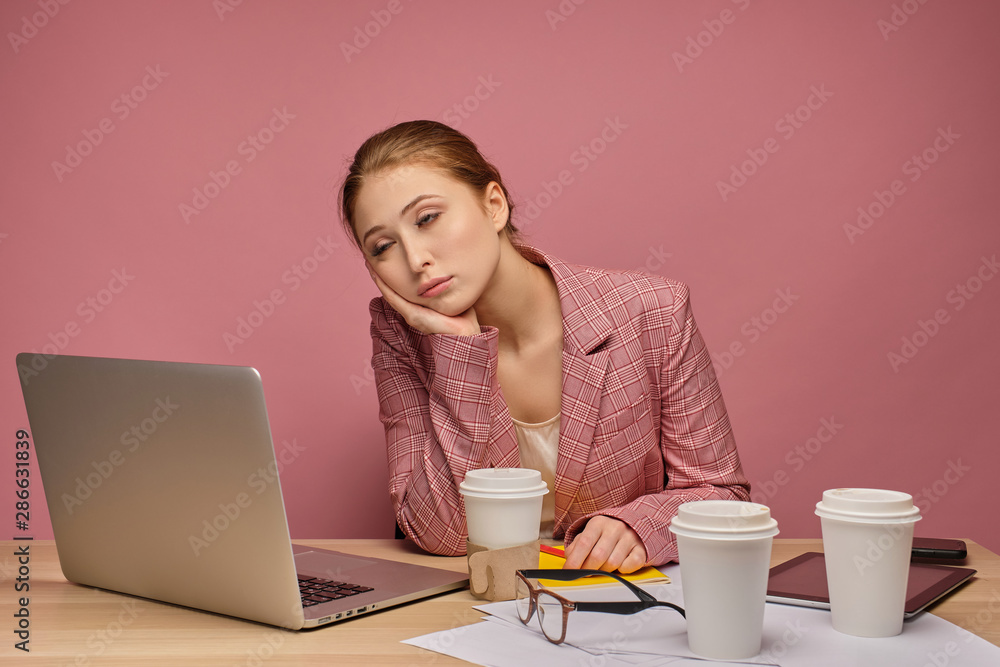 Red-haired girl in a jacket sitting at a table in front of a laptop on a pink background, his head on his hand, falling asleep.
