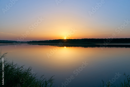 Summer landscape near river in central Russia. The banks of the river with tall grass  bushes and remote forest against setting sun and clear transparent sky. Warm evening sun sets behind the horizon.