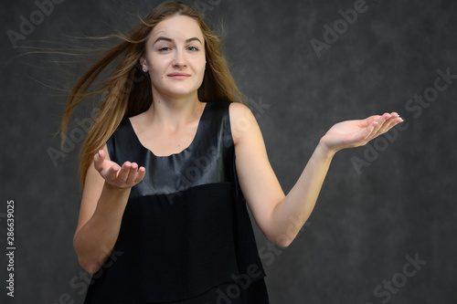 Studio photo portrait of a pretty student girl, brunette young woman with long beautiful hair in a black T-shirt on a gray background. Smiling, talking, showing emotions