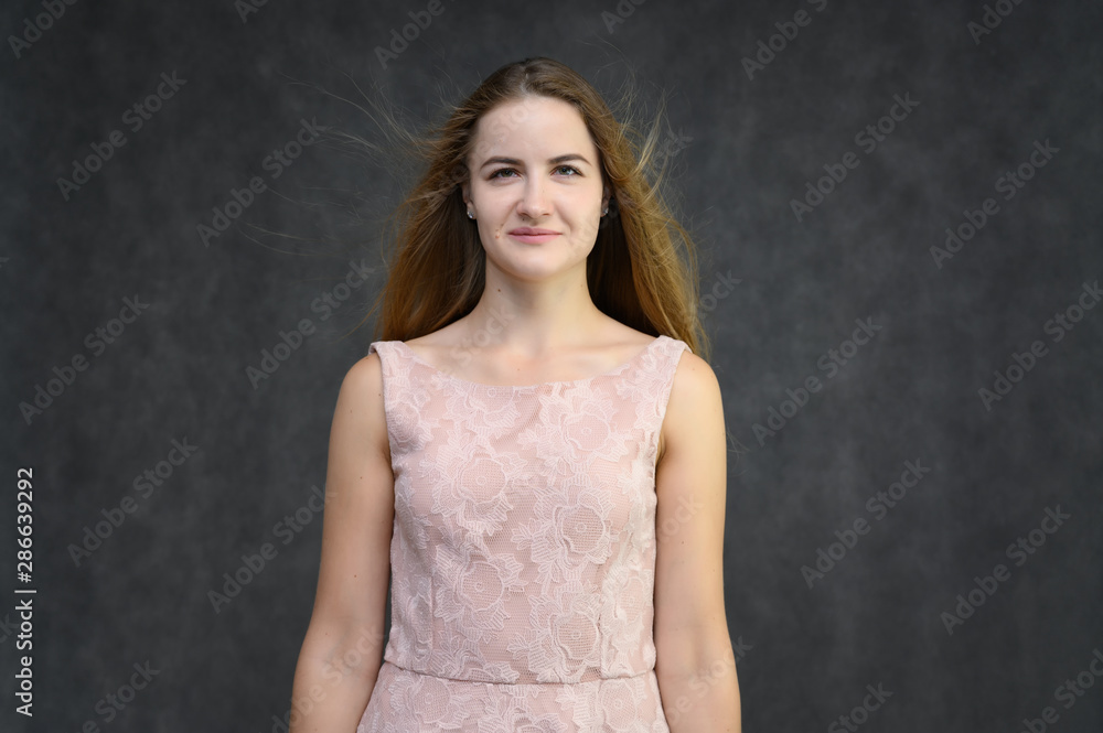 Obraz premium Studio portrait on the waist of a pretty student girl, a young brunette woman with long beautiful hair in a pink dress on a gray background. Smiling, talking, showing emotions