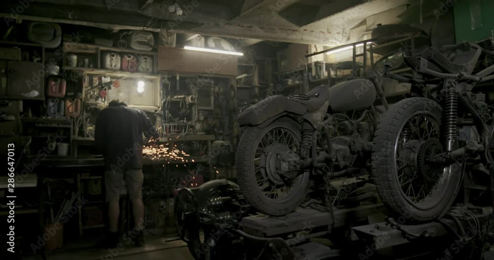 Metalworker working on a grinder with flying fiery sparks in a workshop with rows of stored vintage motorbikes in dim light Metalworker working on a grinder with flying fiery sparks in a workshop with rows of stored vintage motorbikes in dim light