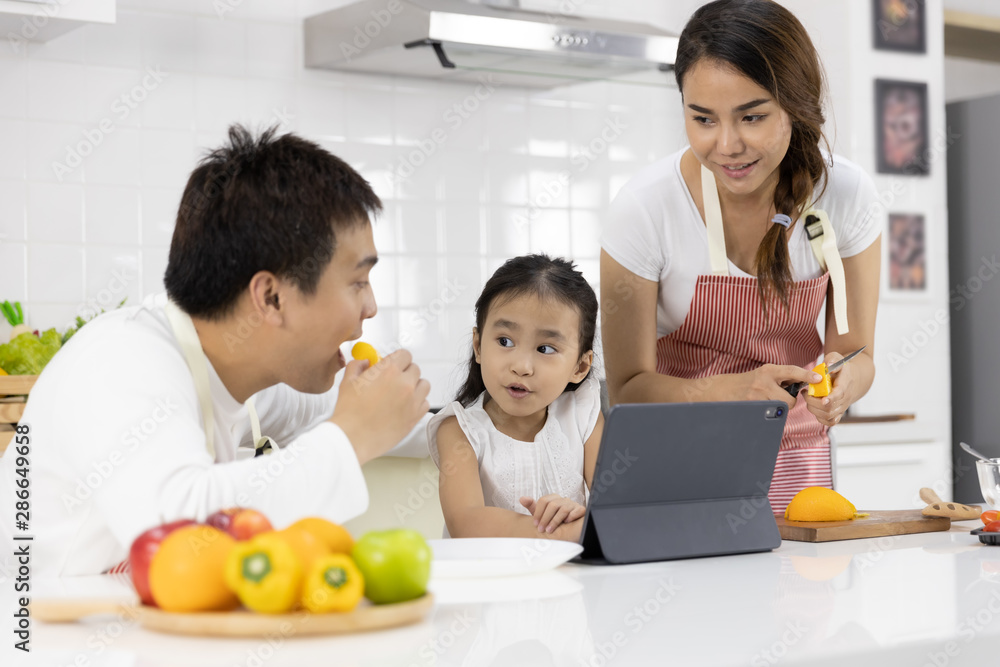 Happy Asian family make a cooking. Father, Mother and Daughter are preparing fruit orange and see Digital tablet in the kitchen at home. Healthy food concept and happy holidays Happy Asian family make a cooking. Father, Mother and Daughter are preparing fruit orange and see Digital tablet in the kitchen at home. Healthy food concept and happy holidays