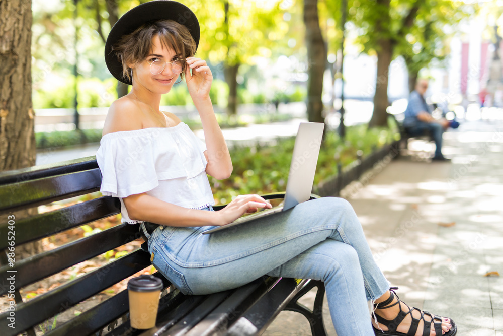 Beautiful woman using laptop while sitting on a bench, drinking takeaway coffee cup Beautiful woman using laptop while sitting on a bench, drinking takeaway coffee cup