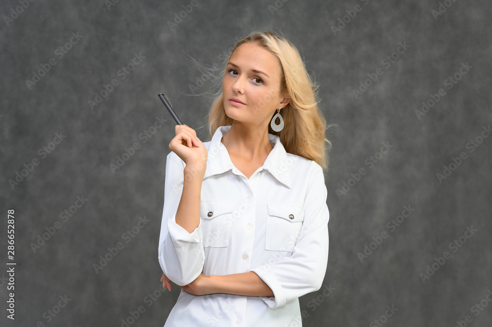 Studio portrait of a pretty blonde student girl, young woman in a white shirt on a gray background. Talking, showing emotions.