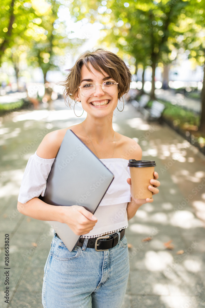Young pretty woman walking with laptop and coffee to go in hand in the park Young pretty woman walking with laptop and coffee to go in hand in the park