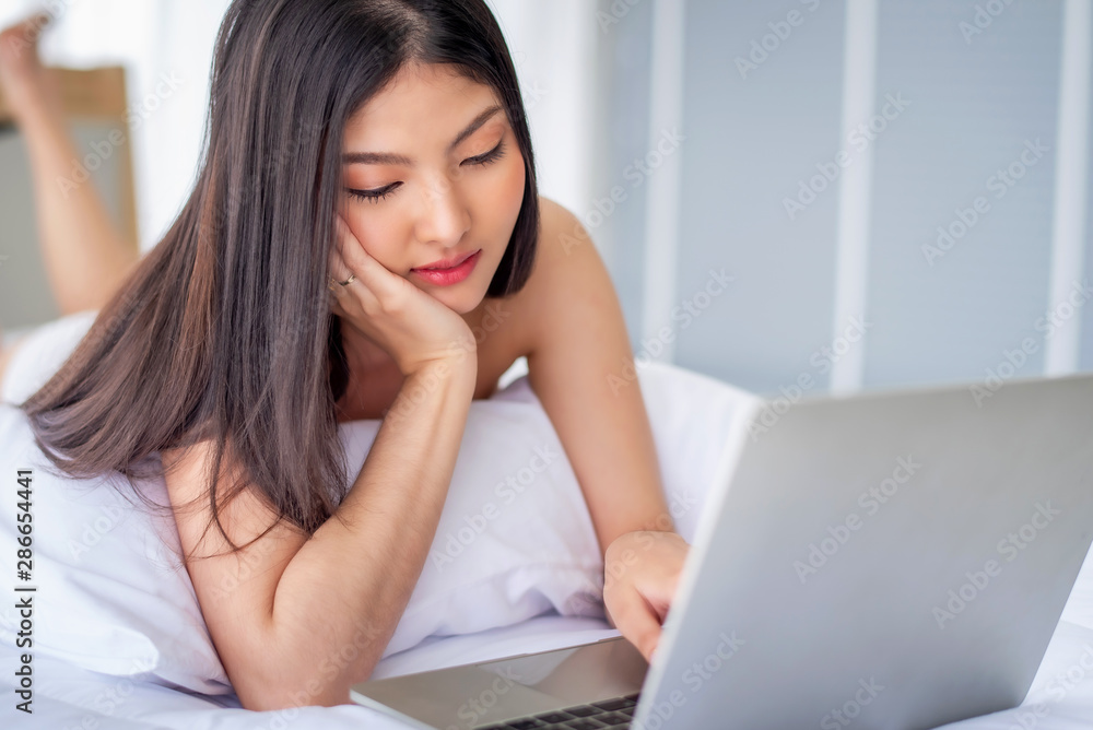 Pretty attractive sensuality young Asian woman using laptop, laying on the white bed at bedroom in the morning Pretty attractive sensuality young Asian woman using laptop, laying on the white bed at bedroom in the morning