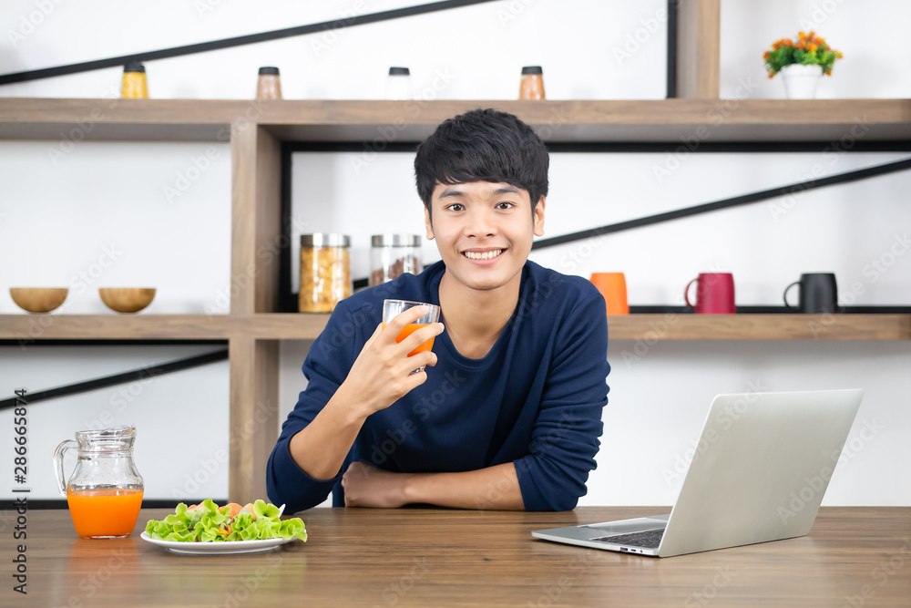 Smiling young Asian businessman working on laptop at office desk and having a fresh vegetable salad for lunch break, hand holding orange juice. Healthy food, eating right and people lifestyle concept. Smiling young Asian businessman working on laptop at office desk and having a fresh vegetable salad for lunch break, hand holding orange juice. Healthy food, eating right and people lifestyle concept.