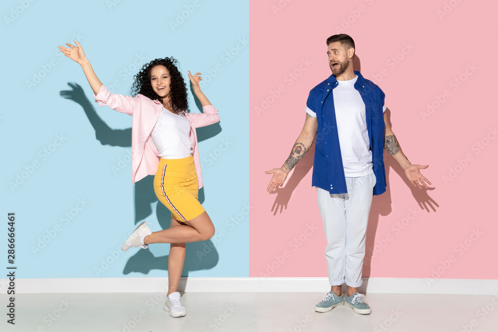 Young emotional caucasian couple in bright casual clothes posing on pink and blue background. Concept of human emotions, facial expession, relations, ad. Man is bored, woman dancing and celebrating. Young emotional caucasian couple in bright casual clothes posing on pink and blue background. Concept of human emotions, facial expession, relations, ad. Man is bored, woman dancing and celebrating.