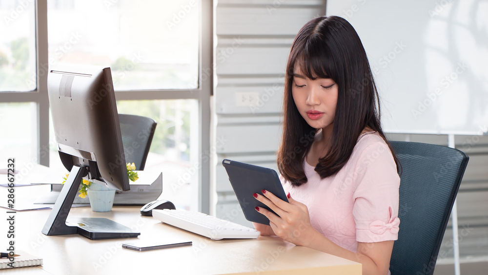 Young Asian business woman sitting and thinking about creative ideas, holding tablet on hand while working at office desk in modern workspace Young Asian business woman sitting and thinking about creative ideas, holding tablet on hand while working at office desk in modern workspace