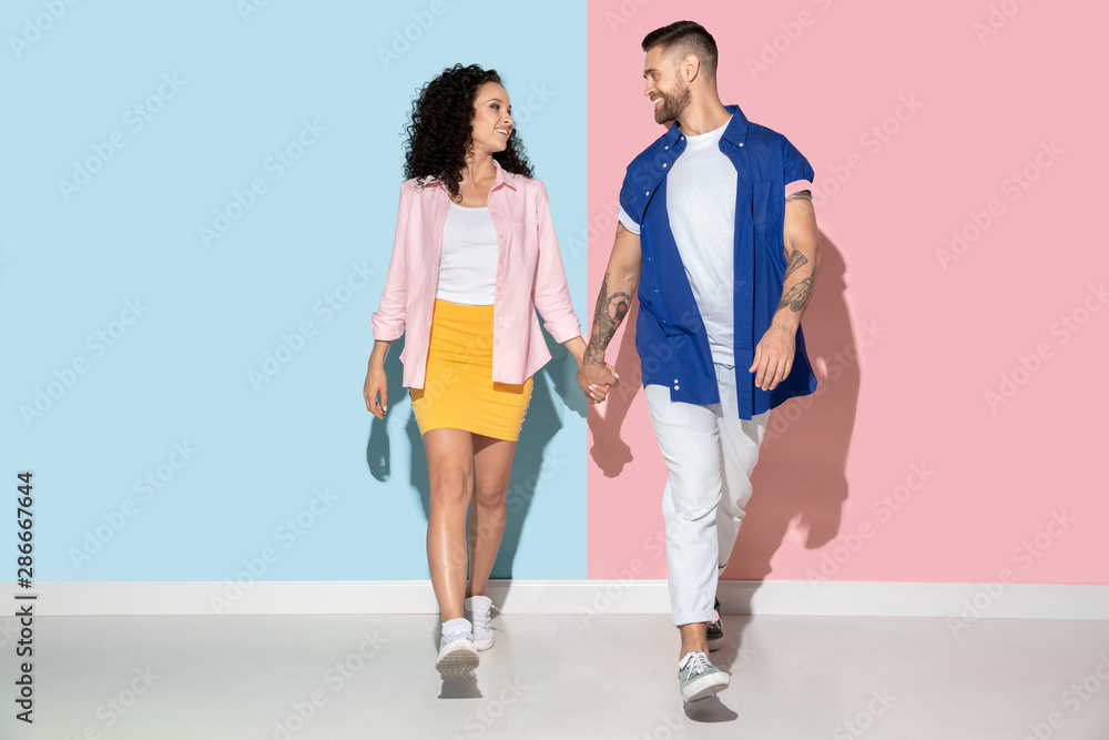 Young emotional caucasian couple in bright clothes posing on pink and blue background. Concept of human emotions, facial expession, relations, ad. Man and woman holding hangs and going, smiling. Young emotional caucasian couple in bright clothes posing on pink and blue background. Concept of human emotions, facial expession, relations, ad. Man and woman holding hangs and going, smiling.