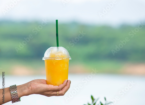 Hand holding a glass of cold Orange juice smoothie Background blurry views tree and water.
