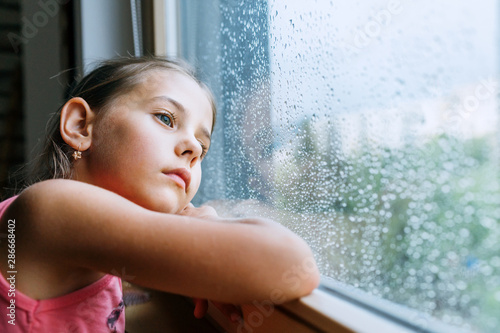 Little sad girl pensive looking through the window glass with a lot of raindrops. Sadness childhood concept image. photo