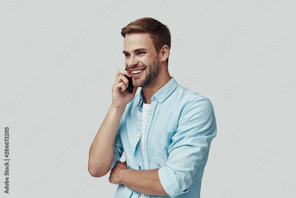 Handsome young man talking on the phone and smiling while standing against grey background Handsome young man talking on the phone and smiling while standing against grey background