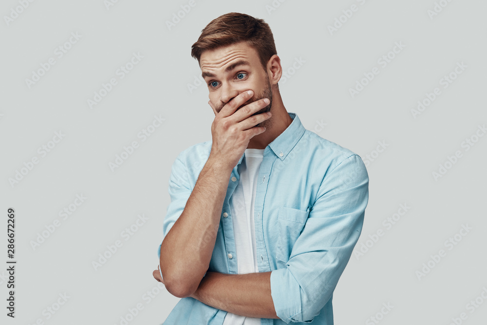 Surprised young man covering mouth with hand while standing against grey background Surprised young man covering mouth with hand while standing against grey background