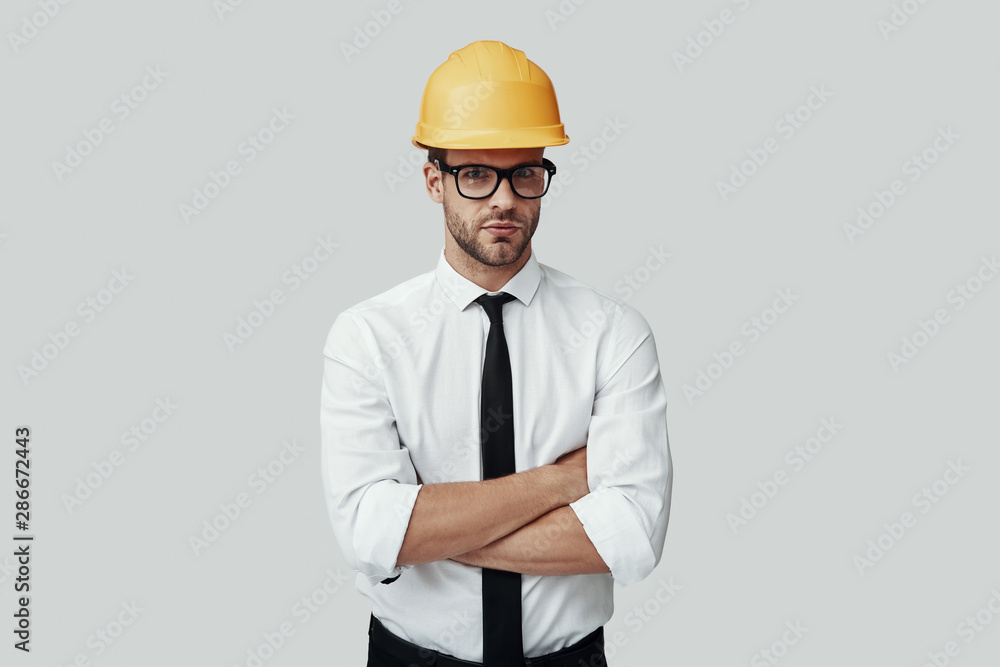 Handsome young man in helmet looking at camera and keeping arms crossed while standing against grey background Handsome young man in helmet looking at camera and keeping arms crossed while standing against grey background