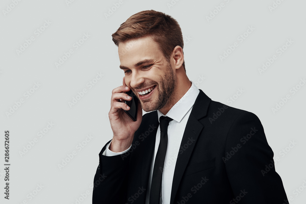 Handsome young man in formalwear talking on the phone and smiling while standing against grey background Handsome young man in formalwear talking on the phone and smiling while standing against grey background