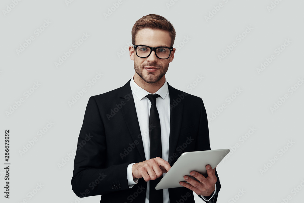 Handsome young man in formalwear working using digital tablet while standing against grey background Handsome young man in formalwear working using digital tablet while standing against grey background