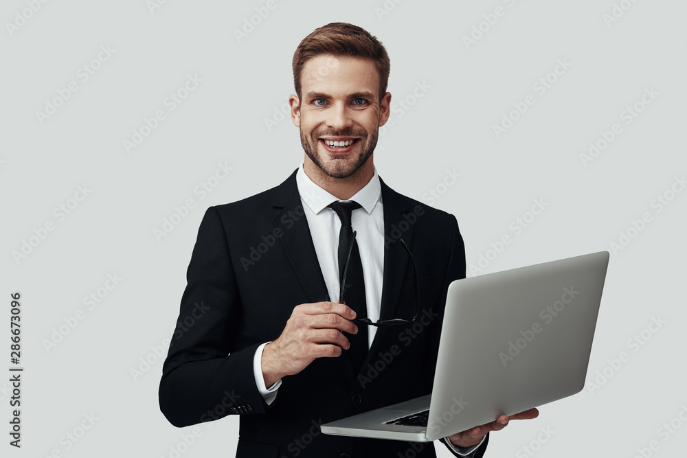 Handsome young man in formalwear working using laptop and smiling while standing against grey background Handsome young man in formalwear working using laptop and smiling while standing against grey background