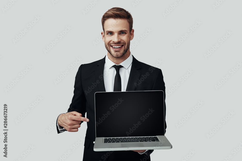 Handsome young man in formalwear showing copy space on laptop while standing against grey background Handsome young man in formalwear showing copy space on laptop while standing against grey background