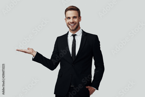 Handsome young man in formalwear looking at camera and pointing copy space while standing against grey background