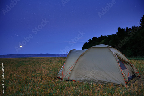 Traveling and camping concept - summer landscape and tent, twilight, moon on dark sky with stars. Beautiful nature - field, forest, plain.