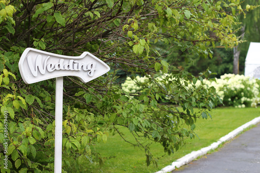 arrow pointer with wedding text on green foliage background. The road leading to the wedding tent. outdoor celebration, field wedding registration on a summer day. Wedding decoration concept arrow pointer with wedding text on green foliage background. The road leading to the wedding tent. outdoor celebration, field wedding registration on a summer day. Wedding decoration concept