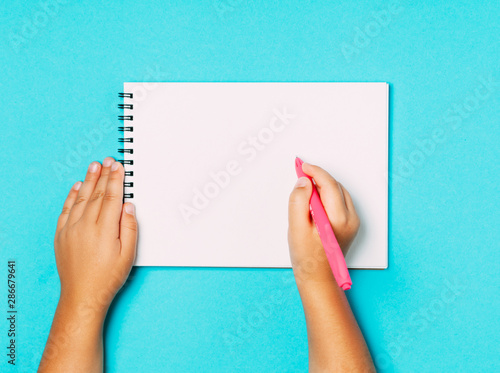 School supplies and stationery with children hand writing in empty notebook on blue table top view.