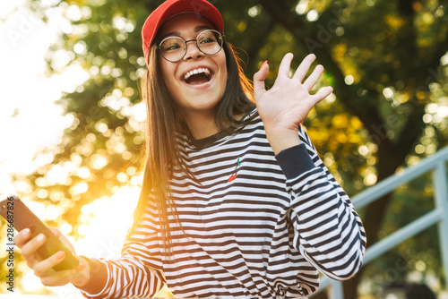 Image of excited caucasian girl waving hand and using cellphone while walking in green park