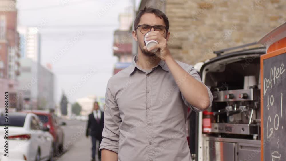 Waist-up portrait arc shot of Caucasian male barista in late twenties standing near mobile van in downtown street, drinking fresh coffee, looking at camera, and business people walking in background Waist-up portrait arc shot of Caucasian male barista in late twenties standing near mobile van in downtown street, drinking fresh coffee, looking at camera, and business people walking in background