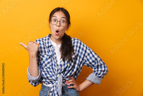 Surprised cute teenage girl in glasses posing isolated over yellow wall background showing copyspace pointing.