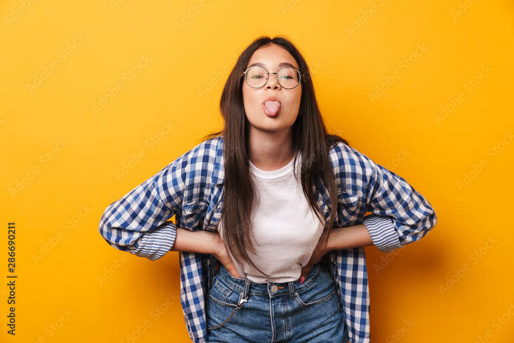 Funny young cute teenage girl in glasses posing isolated over yellow wall background showing tongue. Funny young cute teenage girl in glasses posing isolated over yellow wall background showing tongue.