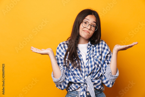 Confused unsured young cute teenage girl in glasses posing isolated over yellow wall background.