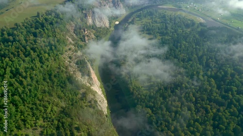Aerial shot from above over the terrain in the fog in the morning. View of the river bend at the foot of the ridge, cliffs and forest valley.