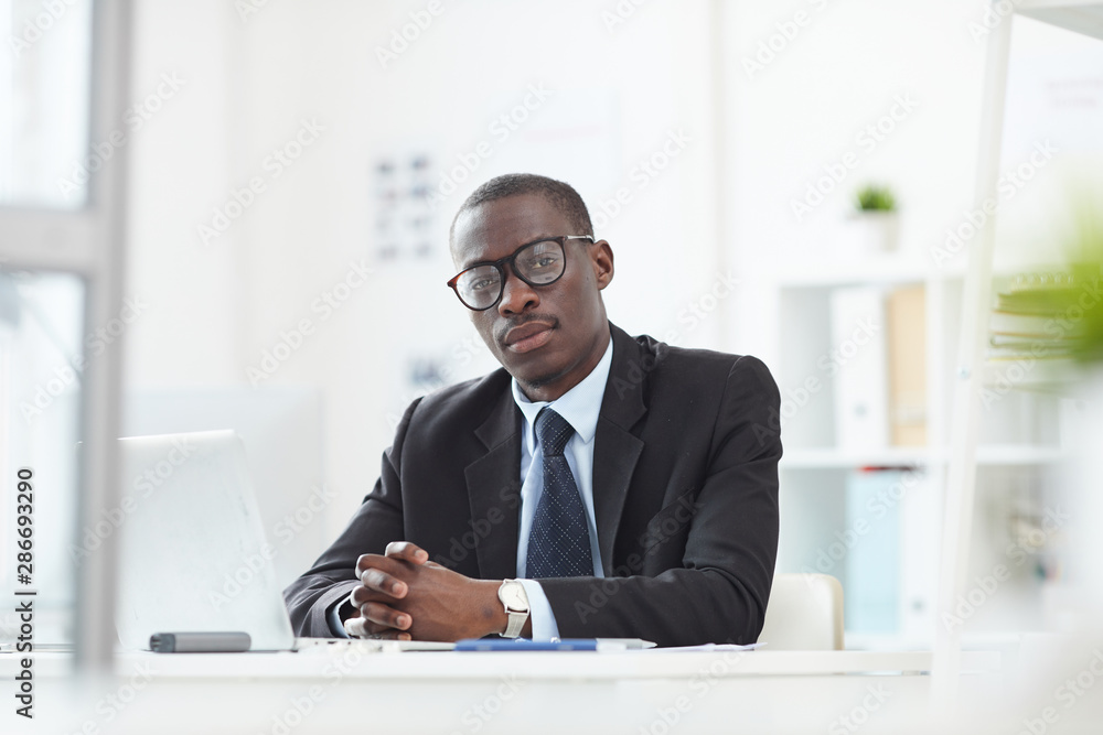 Portrait of serious African male leader in eyeglasses looking at camera while working at the office desk at modern light office